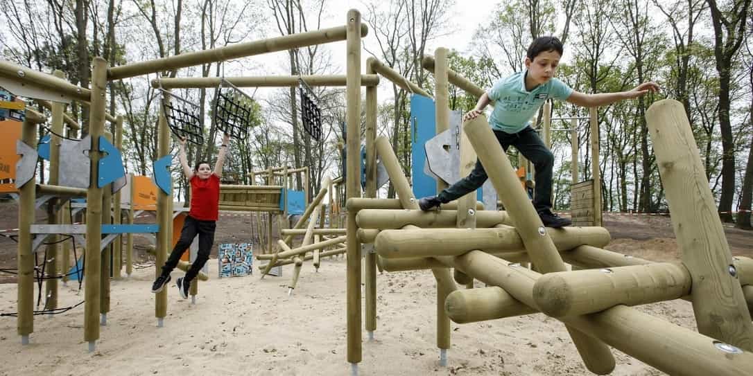 Twee kinderen spelen op een houten klimtoestel in een buitenspeeltuin, waarbij één kind klimt en het andere aan touwen hangt.