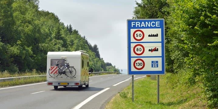 Camper rijdt op een Franse snelweg langs een verkeersbord dat maximumsnelheden aangeeft: 50 km/u binnen de bebouwde kom, 80 km/u op secundaire wegen, en 130 km/u op autosnelwegen. Het bord staat langs een groene, beboste weg onder een blauwe lucht.