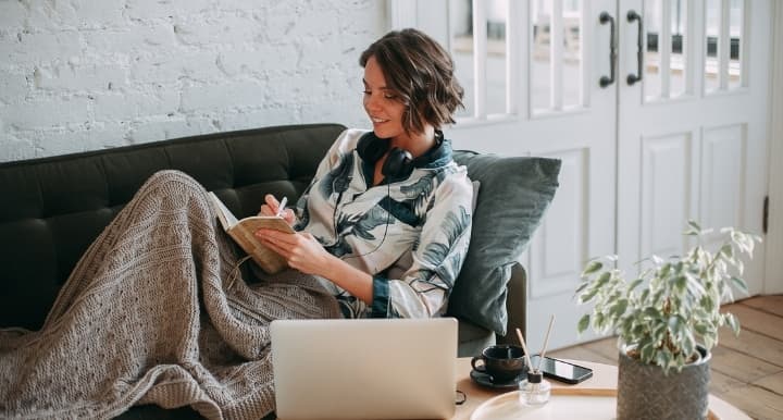 Vrouw zit op de bank onder een deken te schrijven in een boekje met haar koptelefoon op en een laptop en telefoon op de tafel naast haar