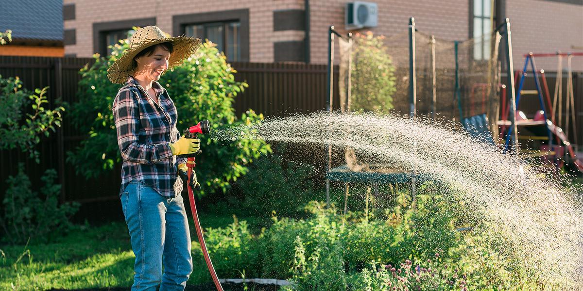Vrouw met strohoed op staat in de tuin met een watersproeier in haar hand de planten nat te sproeien