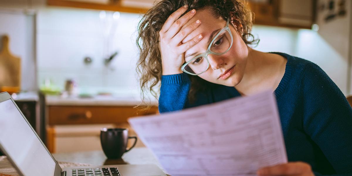 Vrouw met bril aan tafel en ondersteunt haar hoofd met haar hand terwijl ze een laptop voor zich heeft en kijkt naar een papieren document.