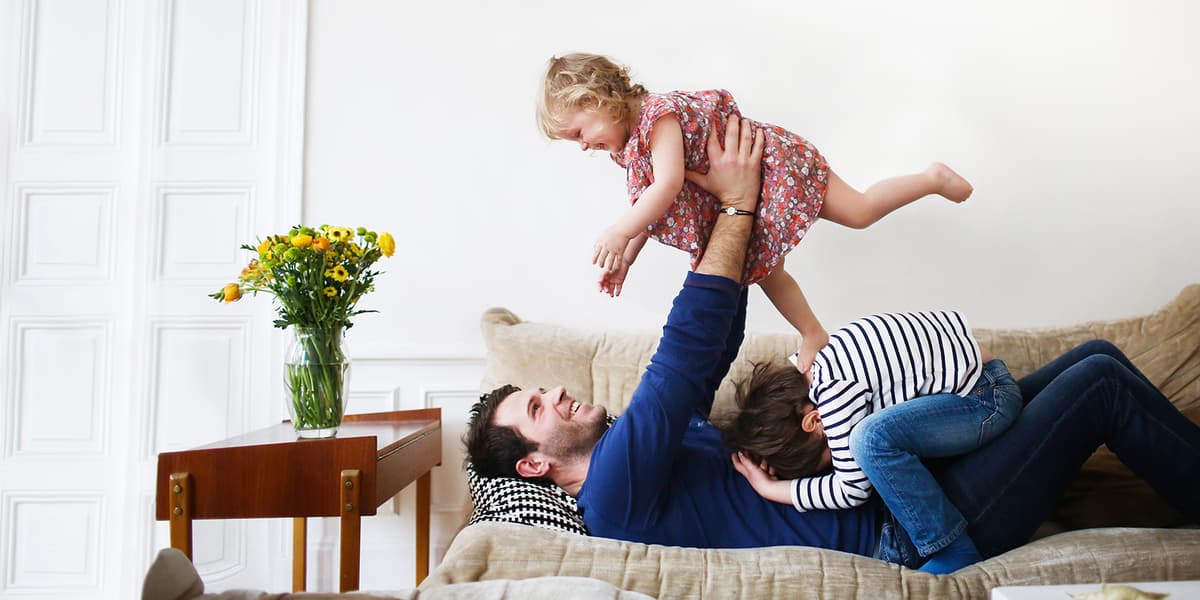 Een jonge vader ligt op de bank en speelt met zijn dochter en zoon. Hij tilt zijn dochter op boven zijn hoofd.