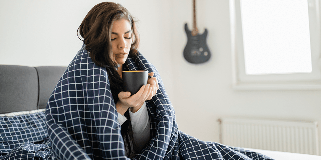 Vrouw zit met een dikke deken om haar heen op bed met een kop koffie in haar hand