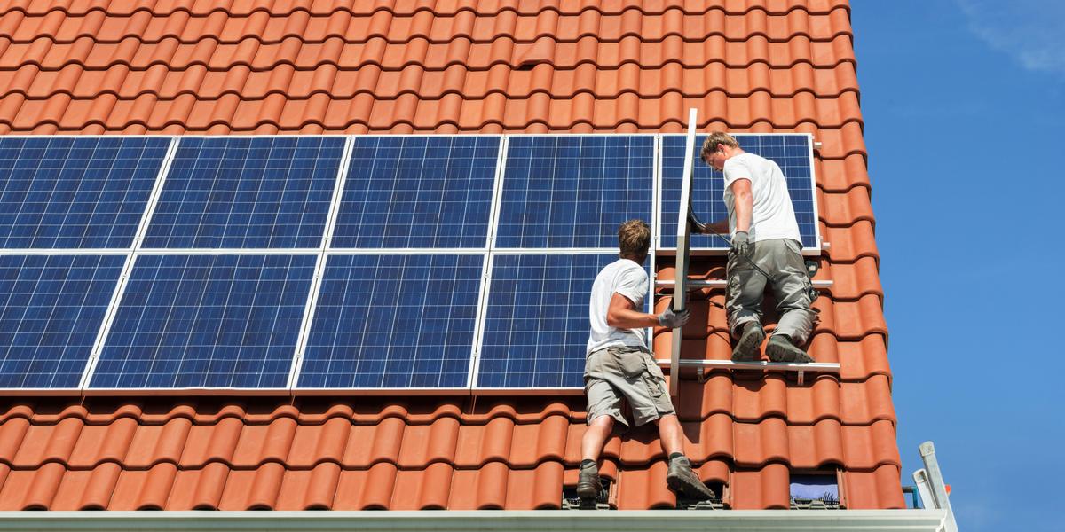 Twee mannelijke installateurs leggen zonnepanelen op het dak van een woning met terracotta dakpannen.