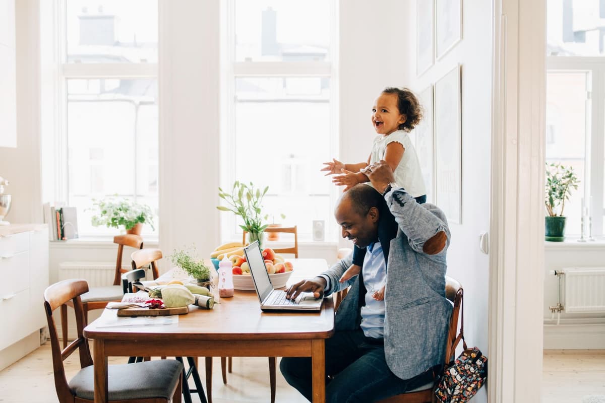 Man zit achter de laptop in de keuken met een kind op zijn schouder