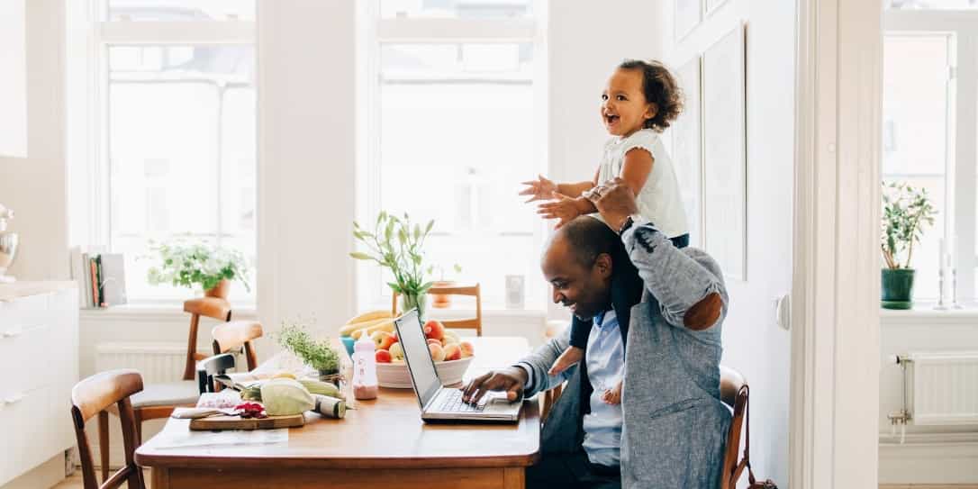 Man zit achter de laptop in de keuken met een kind op zijn schouder