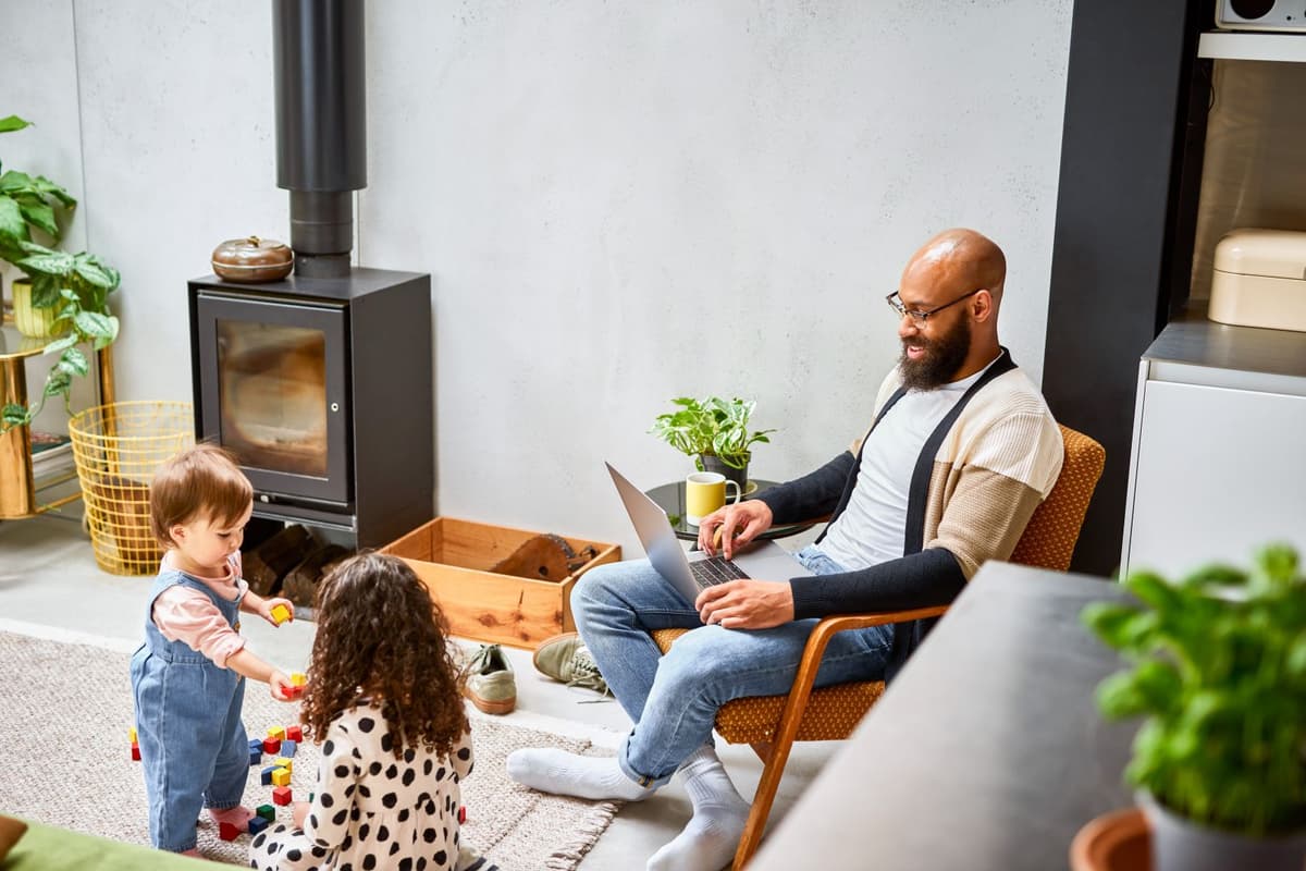 Man zit in de woonkamer met laptop op schoot en twee kinderen spelen op de grond ernaast