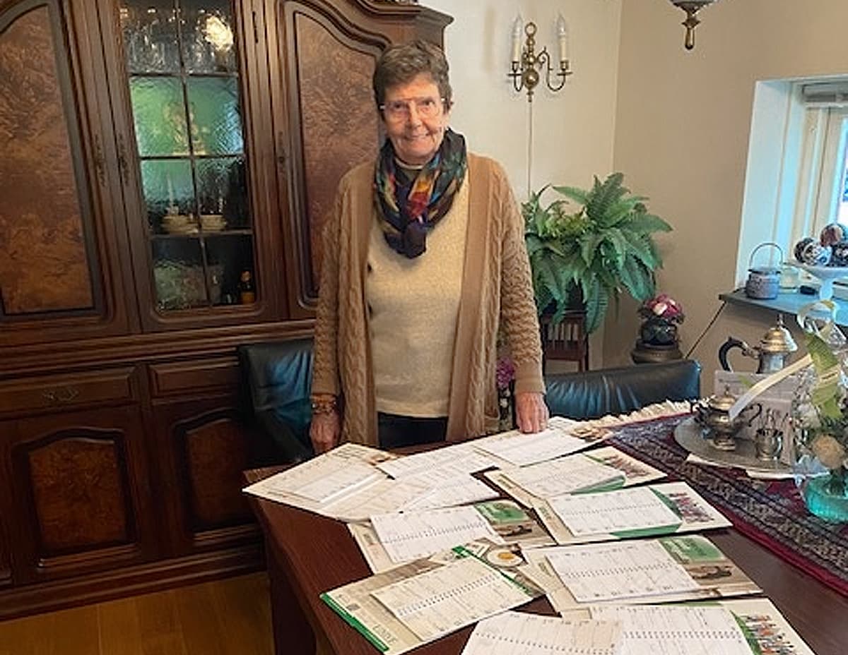 Foto van een glimlachende mevrouw Bakker. Ze staat voor een grote kast en naast een tafel in haar woonkamer. Op tafel liggen allerlei Univé kalenders kriskras verpreid.