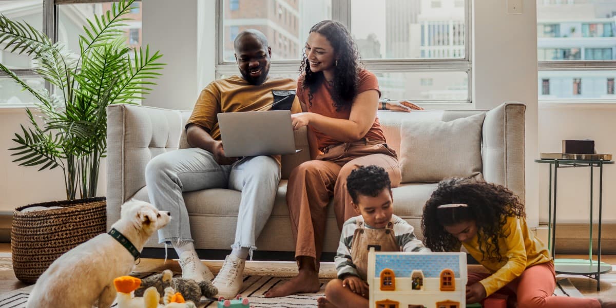 Gezin en hond in een woonkamer. Ouders op de bank kijken lachend naar een laptop, twee kinderen spelen op de grond.