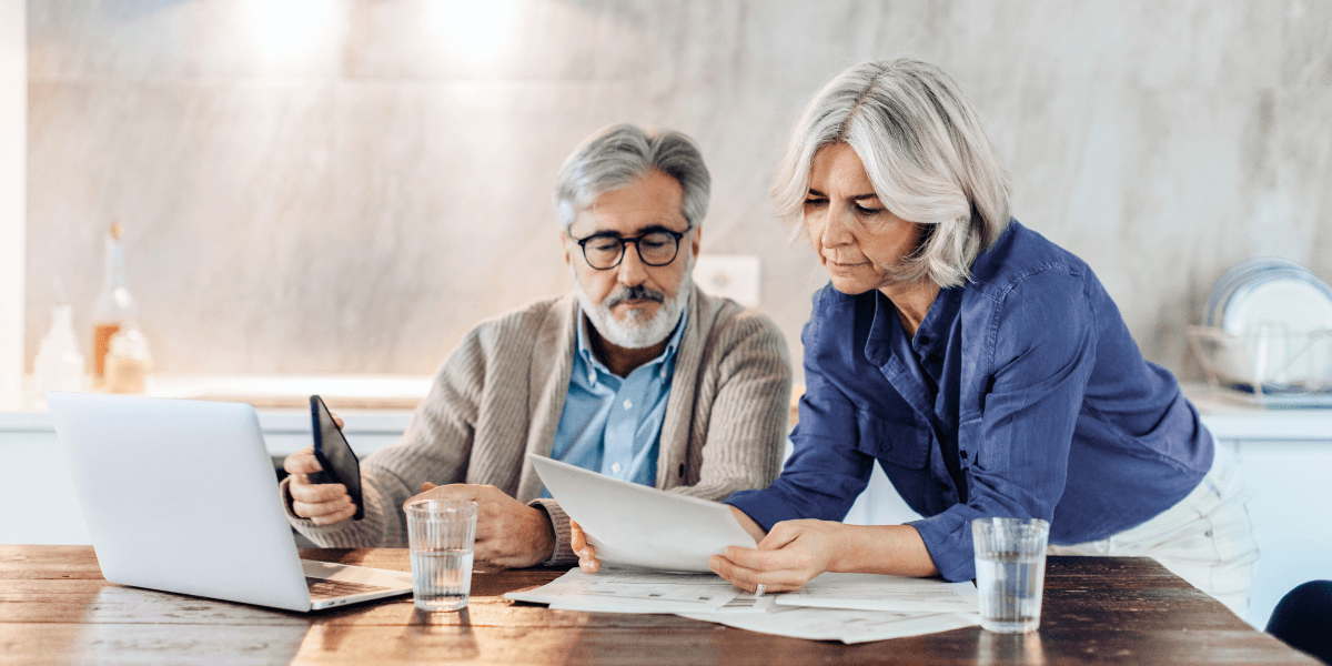 Oude man en vrouw zijn aan de eettafel samen bezig op hun laptop. Ze kijken naar papieren documenten en de man heeft een telefoon in zijn hand.
