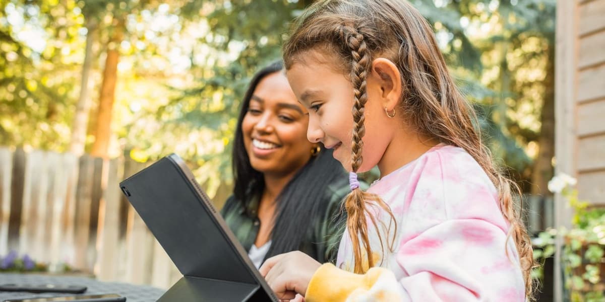Jong meisje zit in de tuin, lachend naar een iPad. Naast haar zit een jonge vrouw.