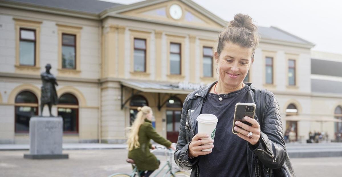 Vrouw loopt bij een station met een koffiebeker in de hand. In haar andere hand houdt ze haar iPhone vast. Ze lacht naar het scherm.