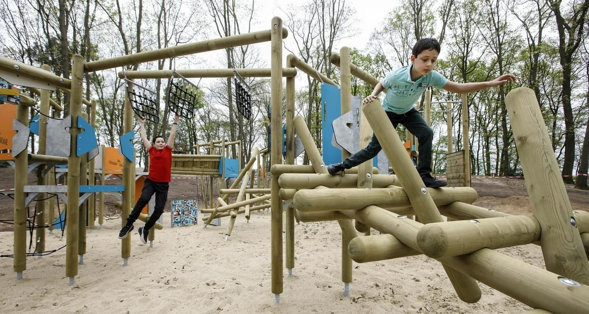 Twee kinderen spelen op een houten klimtoestel in een buitenspeeltuin, waarbij één kind klimt en het andere aan touwen hangt.