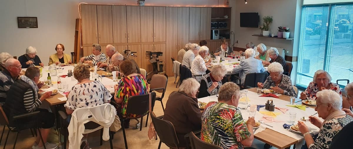 Een groep oudere mensen zit aan drie grote tafels in een lichte, gezellige zaal van wijkcentrum Wippolder in Delft. Ze genieten samen van een warme maaltijd, met aandacht voor de presentatie van de tafels en servetten. Vrijwilligers zorgen voor het eten en de sfeer, waarbij samenzijn en het bestrijden van eenzaamheid centraal staan. De sfeer is warm en uitnodigend, met veel betrokkenheid en gezelligheid.