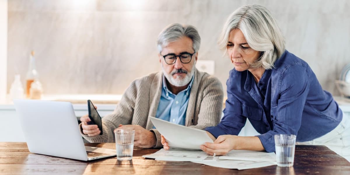 Man en vrouw op leeftijd regelen zaken op laptop terwijl ze kijken naar stapels papier