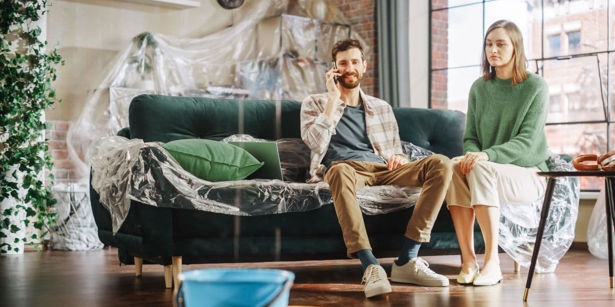 Jong stel zit op bank terwijl ze kijken naar de lekkage in hun woning. Op de vloer staan emmers om het lekkende water op te vangen.