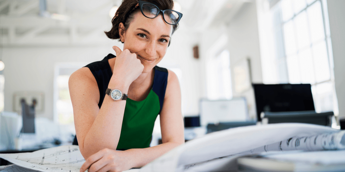 Een vrouwelijke architect zit glimlachend aan een tafel met om zich heen een aantal bouwtekeningen.