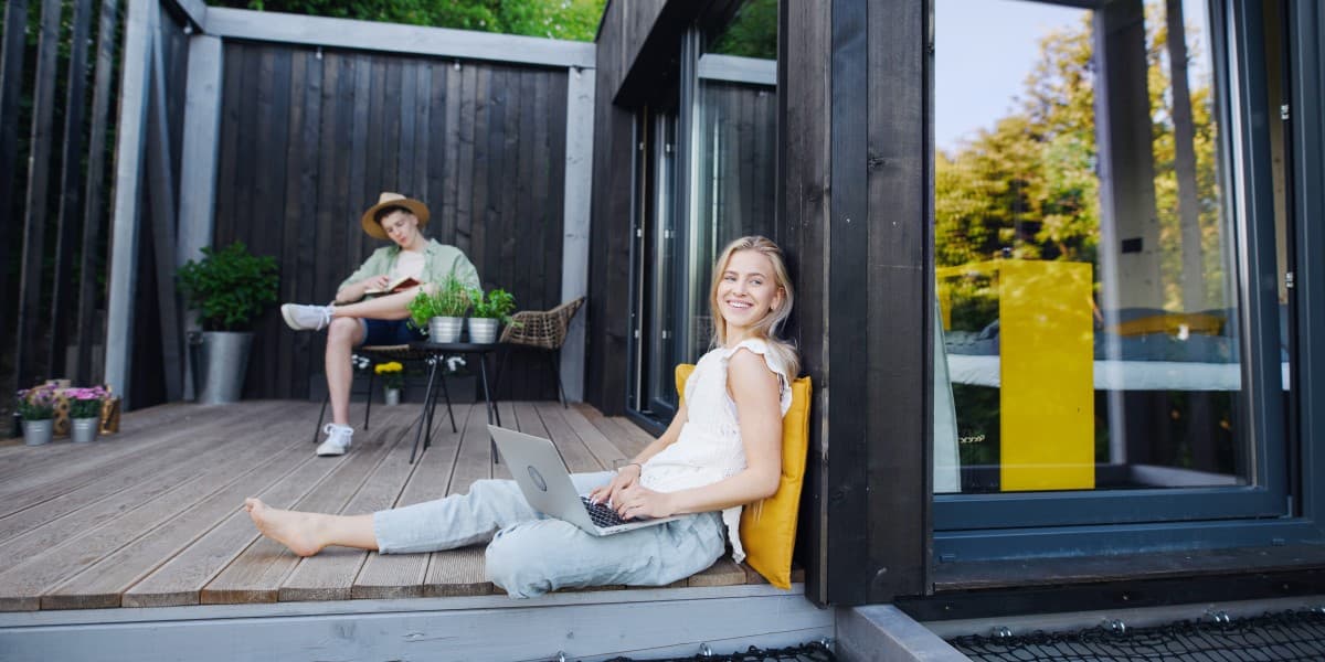 Vrouw zit met een laptop op de grond voor haar recreatiewoning en een man op de achtergrond te lezen