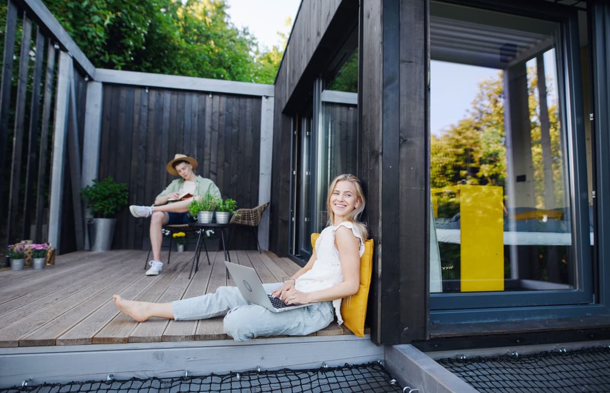 Jonge man en vrouw zitten op de veranda van een houten chalet. De jongen leest een boek en de vrouw heeft haar laptop op schoot.
