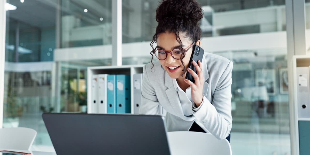 Een vrouw telefoneert terwijl ze lachend op haar laptop kijkt. Op de achtergrond staat een vakkenkast met daarin een aantal ordners.