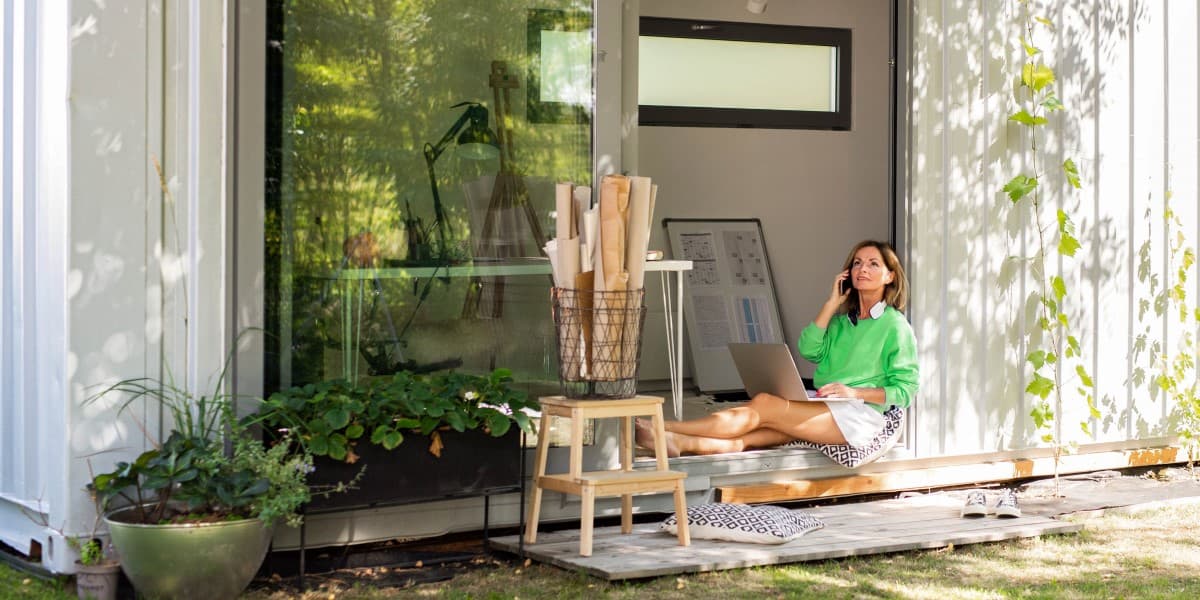 Vrouw zit in de open deur van haar containerwoning met een laptop op schoot te bellen