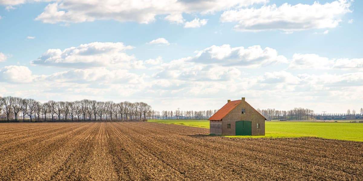 Losse schuur op omgeploegde grond. Blauwe, bewolkte lucht boven de schuur en bomen op de achtergrond.