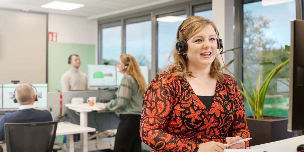 Medewerker belt met headset op, drie collega's overleggen op de achtergrond