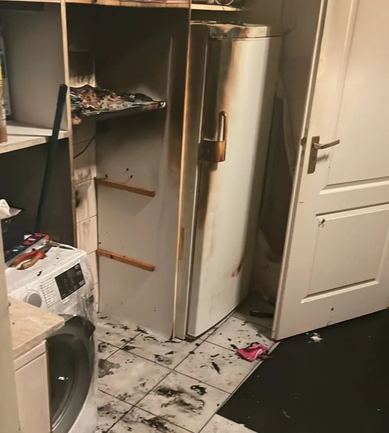 Bijkeuken na drogerbrand, met zwartgeblakerde koelkastdeur, verbrande planken, roetsporen op muren en een witte tegelvloer vol asresten.
