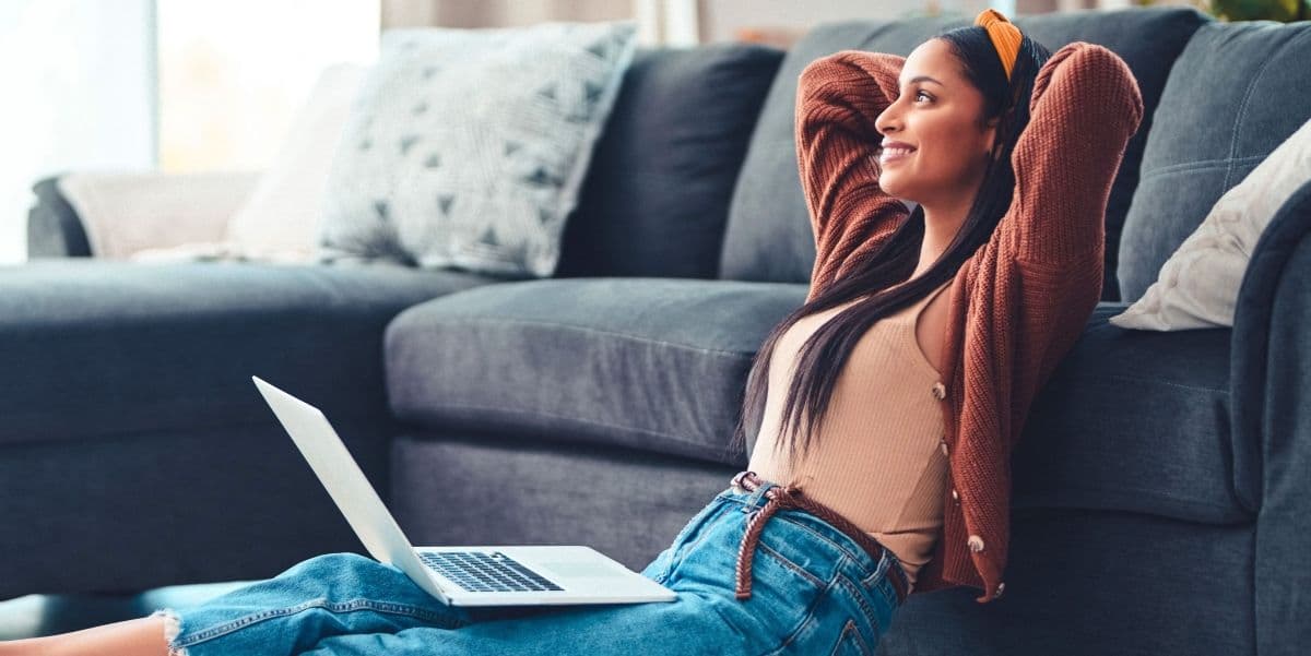 Jonge vrouw met laptop op schoot zit relaxt en tevreden tegen de bank