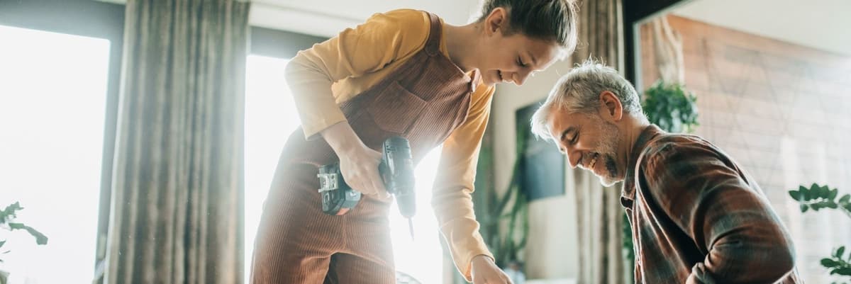 Man met grijs haar en meisje in bruin overall en geel shirt zijn aan het klussen in huis. Het meisje heeft een boormachine in haar hand. Ze lachen allebei.