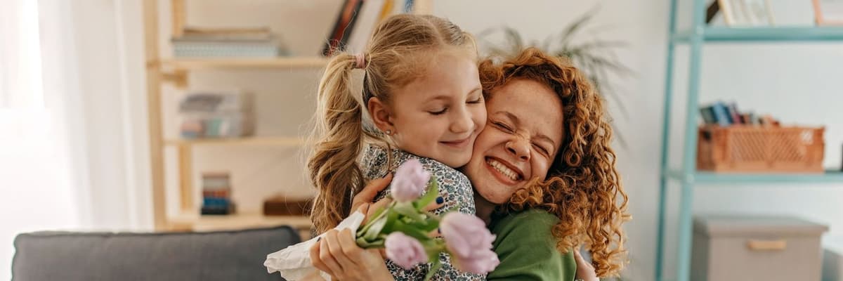 Moeder met rode krullen en groene trui knuffelt haar jonge dochter. De moeder houdt een bos roze tulpen in haar hand.