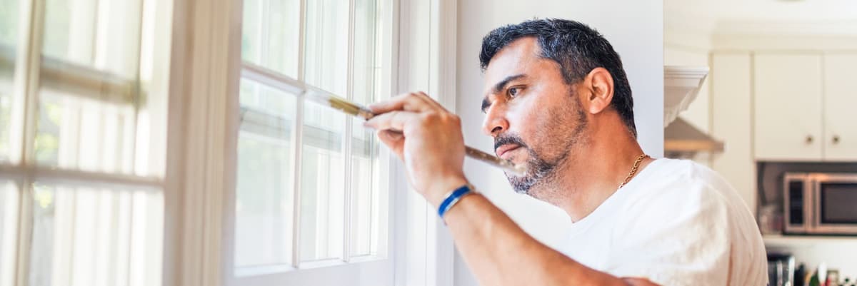 Man met zwart haar en baard verft zijn kozijnen met witte verf.
