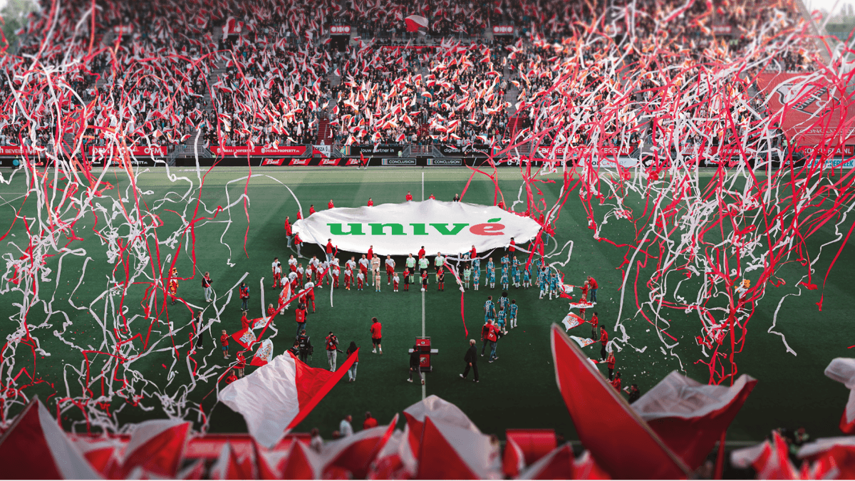 Stadion Galgenwaard vol met supporters die vlaggen zwaaien, met op de middenstop een doek met Univé-logo