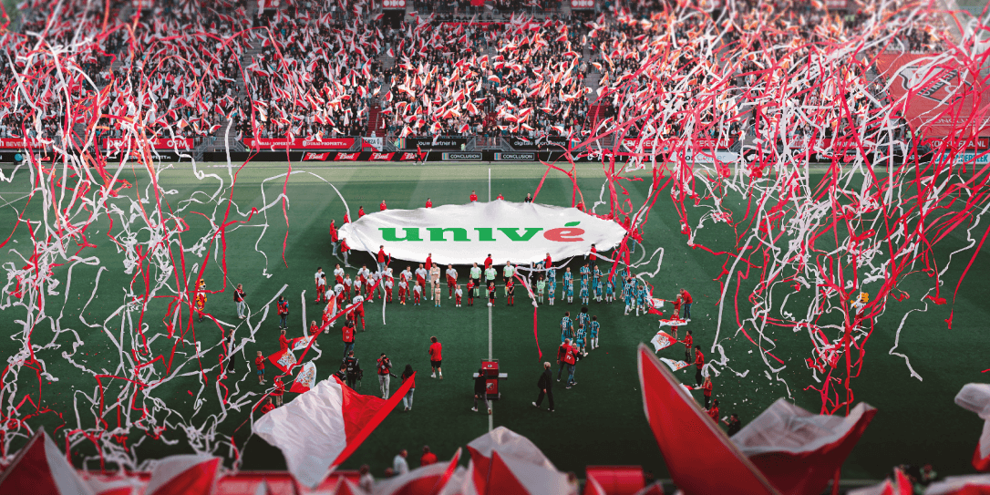 Stadion Galgenwaard vol met supporters die vlaggen zwaaien, met op de middenstop een doek met Univé-logo