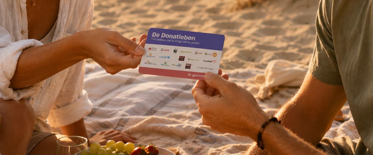 Een persoon geeft op het strand tijdens een picknick een cadeaukaart met de tekst “De Donatiebon – Het cadeau dat je krijgt om te geven” aan iemand anders.