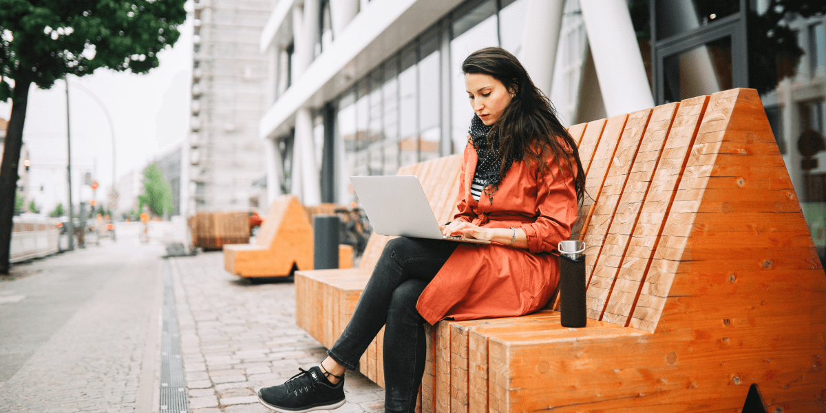 Vrouw zit op een bankje buiten haar kantoor en werkt op haar laptop